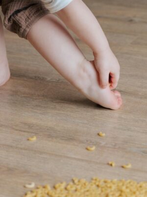 Crop faceless toddler standing barefoot on floor and trying to remove stuck pasta from foot while playing and developing fine motor skills at home